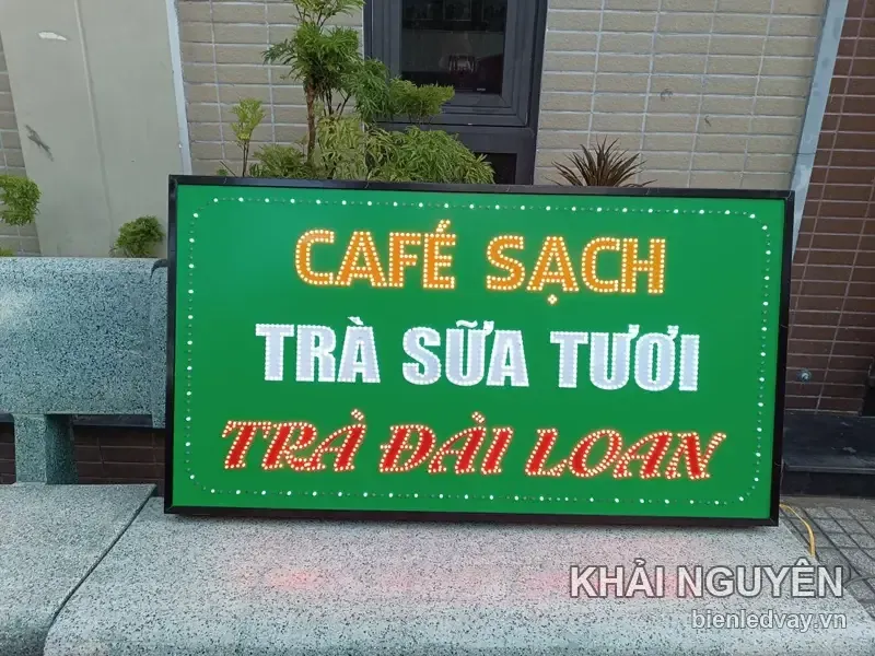 Biển vẫy nền xanh lá với ba dòng chữ: CAFÉ SẠCH vàng, TRÀ SỮA TƯƠI trắng, TRÀ ĐÀI LOAN đỏ, dựng trên bệ đá ngoài sân