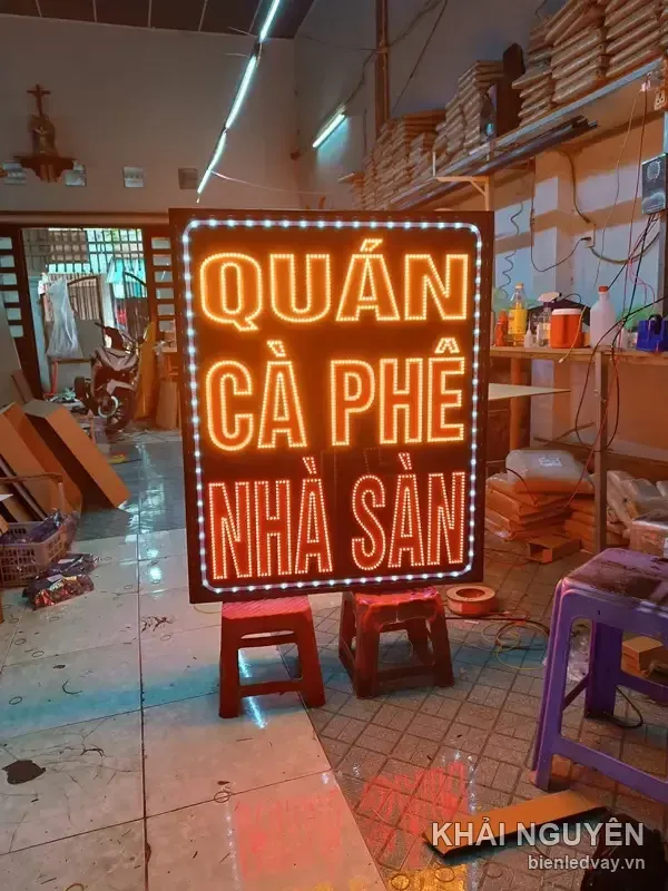 Biển vẫy LED khổ lớn có chữ QUÁN CÀ PHÊ NHÀ SÀN màu cam trên nền đen, viền LED bóng tròn vàng, đặt trên ghế nhựa ở xưởng