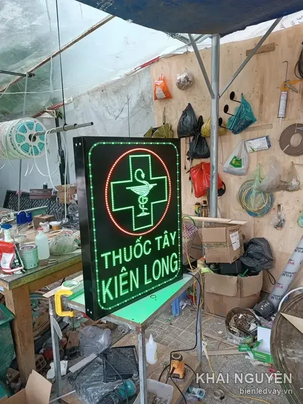 Thuốc Tây Kiên Long với chữ thập dược xanh lá trong vòng tròn LED đỏ, chữ xanh lá trên nền đen, dựng dưới mái bạt