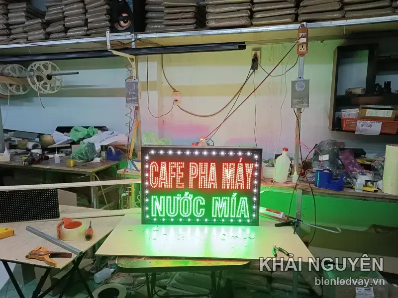 Hình ảnh toàn cảnh xưởng chụp biển vẫy CAFE PHA MÁY chữ đỏ và NƯỚC MÍA xanh lá, phía sau có kệ chất bao xi măng.