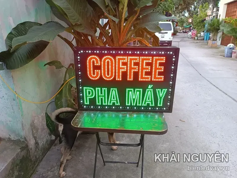Biển vẫy COFFEE PHA MÁY nền đen viền LED bóng tròn, chữ màu đỏ và xanh lá, kê trên ghế xếp ngoài hẻm.