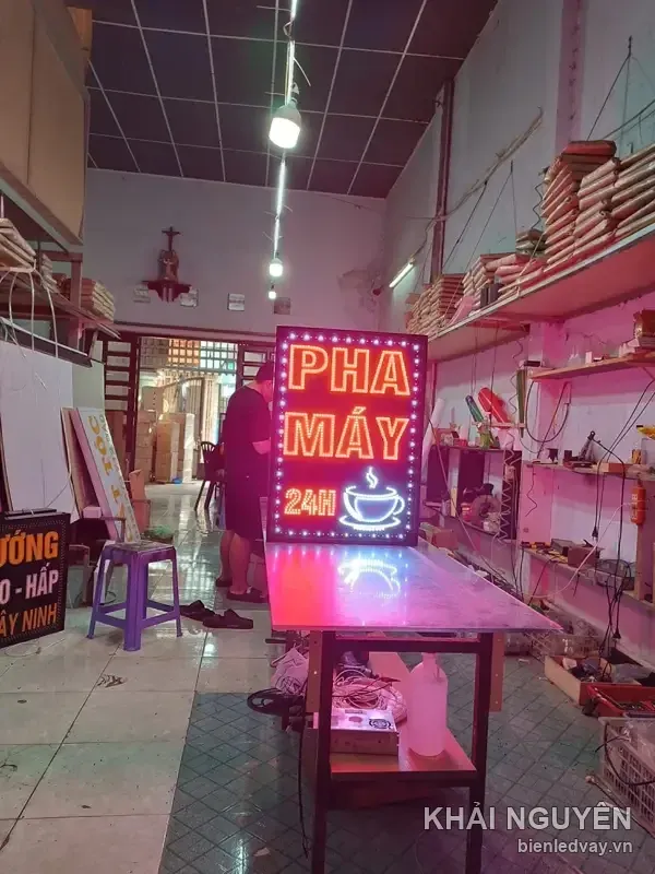 Hình biển PHA MÁY 24H nền đen viền bóng LED trắng, có chữ LED cam và hình tách cà phê bốc khói, đang thử sáng ở xưởng.
