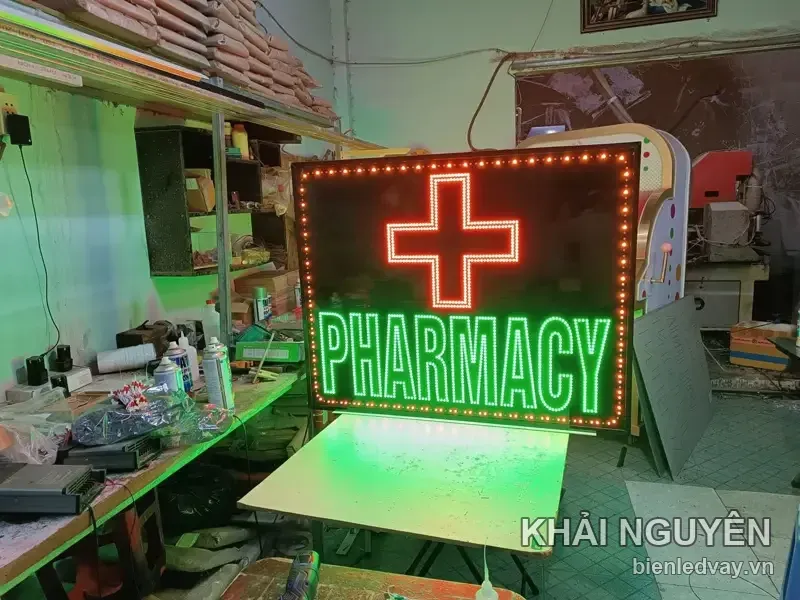 Biển vẫy LED PHARMACY chụp chếch trong xưởng, viền bóng tròn cam nhấp nháy, ánh xanh hắt xuống mặt bàn