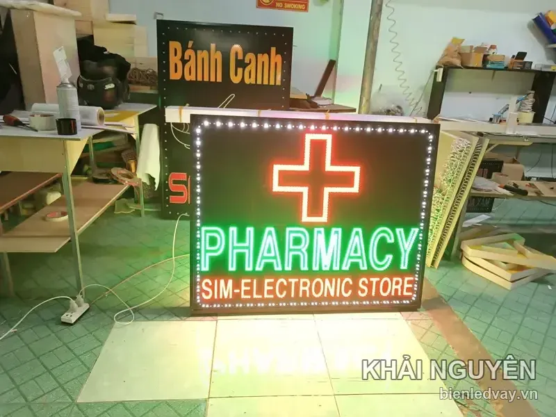 Biển vẫy Pharmacy Sim-Electronic Store có nền đen, thập giá đỏ, chữ LED xanh lá và viền bóng trắng.