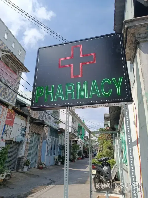 Biển vẫy PHARMACY treo trước cửa trong hẻm, có chữ LED xanh lá, chữ thập viền đỏ trên nền đen và viền LED trắng