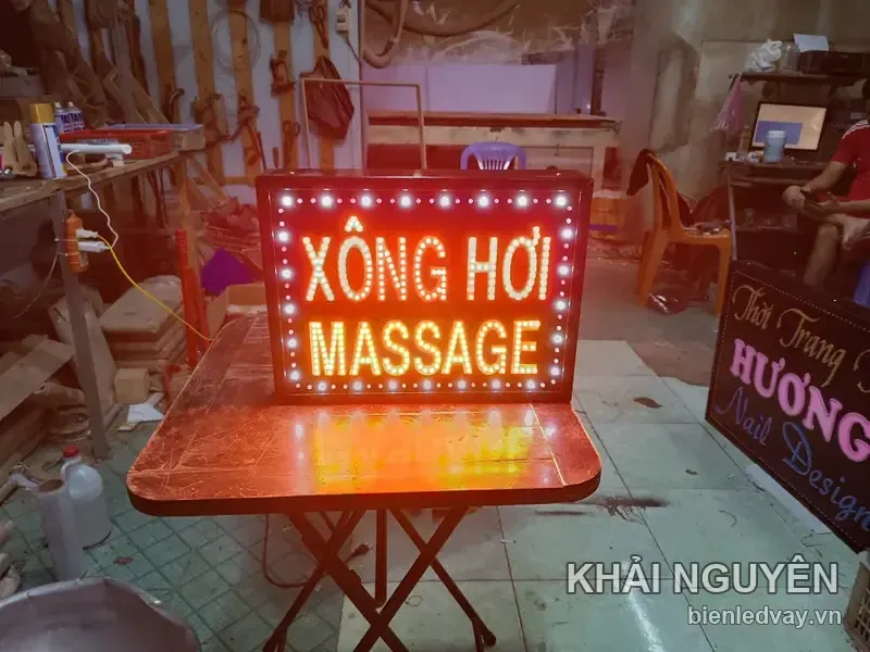 Biển vẫy XÔNG HƠI chữ đỏ, MASSAGE chữ vàng, viền bóng LED trắng, đặt trên bàn xếp mặt gỗ đỏ trong xưởng