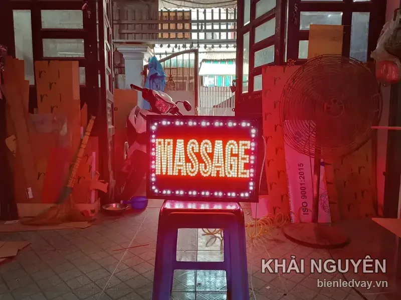 Biển vẫy chữ MASSAGE bóng LED đỏ cam trên nền đen, viền bóng tròn trắng, dựng trên ghế nhựa ngay cửa xưởng