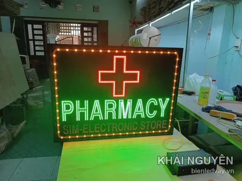 Biển vẫy PHARMACY viền bóng vàng, thập giá đỏ, chữ xanh lá hắt vệt sáng xanh xuống nền gạch xưởng
