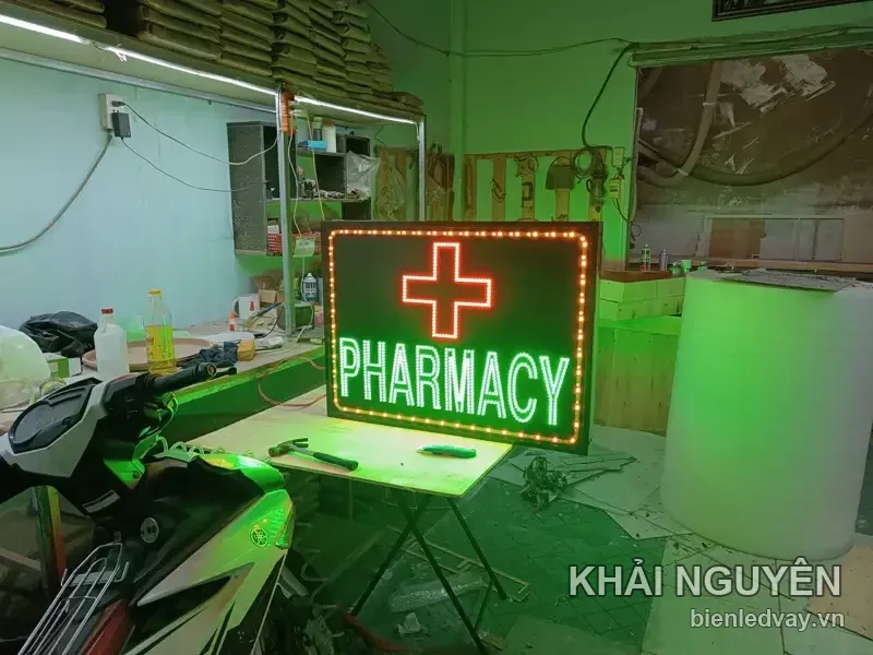 Biển vẫy PHARMACY chữ LED xanh lá, chữ thập viền đỏ, viền bóng tròn vàng, dựng trên bàn gấp giữa xưởng