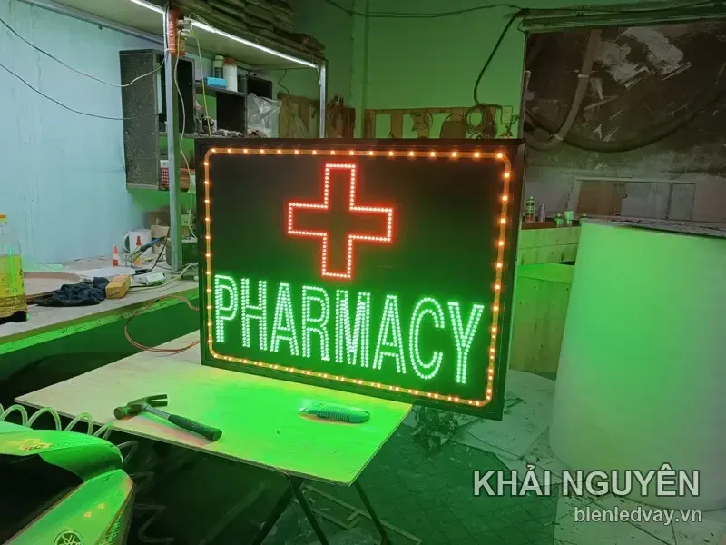 Biển vẫy nhà thuốc chữ thập LED đỏ và chữ PHARMACY xanh lá, viền LED vàng, đặt trên bàn gỗ trong xưởng