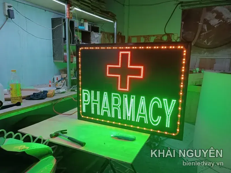 Biển vẫy PHARMACY chụp chếch trái, chữ thập LED đỏ nổi trên nền đen, chữ xanh lá, viền bóng LED vàng cam