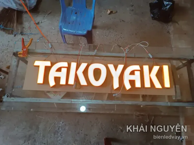 Chữ nổi mica TAKOYAKI gắn trên nền gỗ, sáng đèn vàng ấm, đang được đấu dây chạy thử tại xưởng
