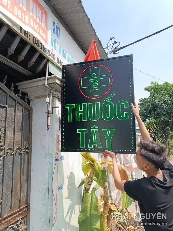 Thợ đưa biển vẫy LED THUỐC TÂY nền đen chữ xanh lên tường nhà, chụp ngoài hẻm nắng buổi trưa
