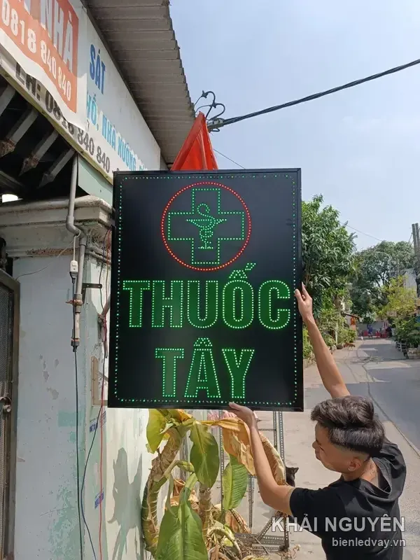 Biển vẫy LED THUỐC TÂY nền đen chữ xanh lá đang được lắp lên mặt tiền, phía sau là hẻm nhỏ nhiều cây xanh