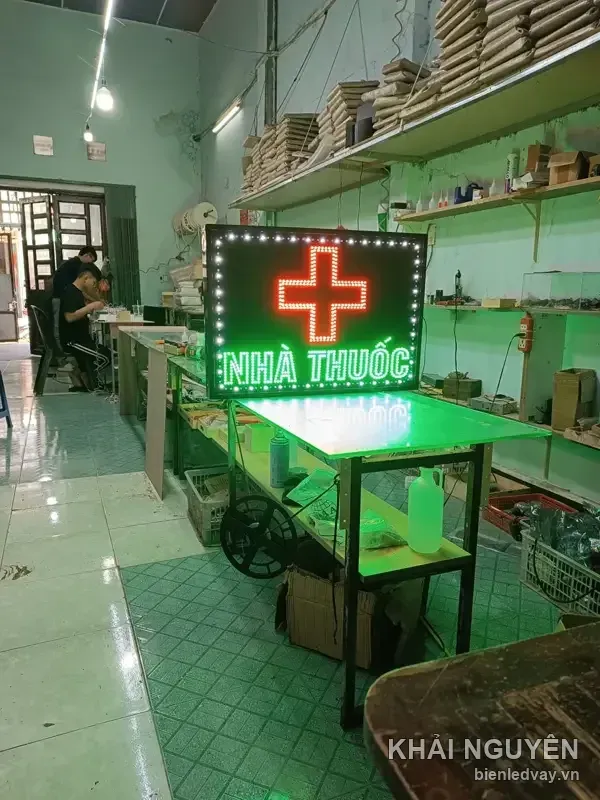 Ảnh toàn cảnh xưởng có biển vẫy NHÀ THUỐC chữ xanh lá, chữ thập đỏ, viền bóng trắng, hắt sáng xanh lên mặt bàn