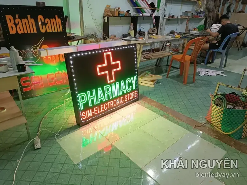 Biển vẫy PHARMACY chữ thập đỏ, dòng SIM-ELECTRONIC STORE chữ đỏ, viền LED trắng, dựng trên nền gạch xưởng