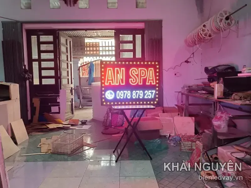 Biển vẫy AN SPA chữ đỏ kèm số liên hệ trên biển, viền LED bóng vàng, dựng trên chân đế chữ X trong xưởng