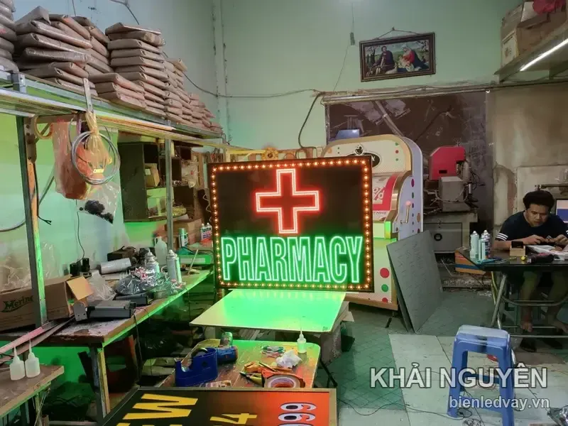 Biển vẫy LED PHARMACY đang thử sáng ở góc xưởng, chữ thập đỏ, chữ xanh, phía sau có thợ ngồi làm việc