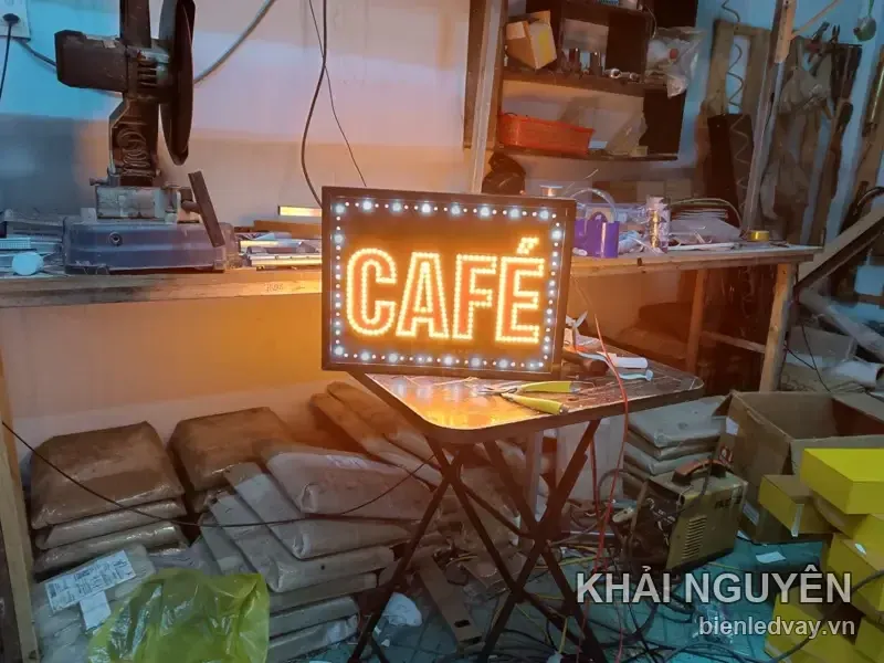 Biển vẫy nền đen viền bóng tròn trắng, có chữ CAFÉ bằng LED vàng, đang dựng trên bàn gấp ở xưởng.