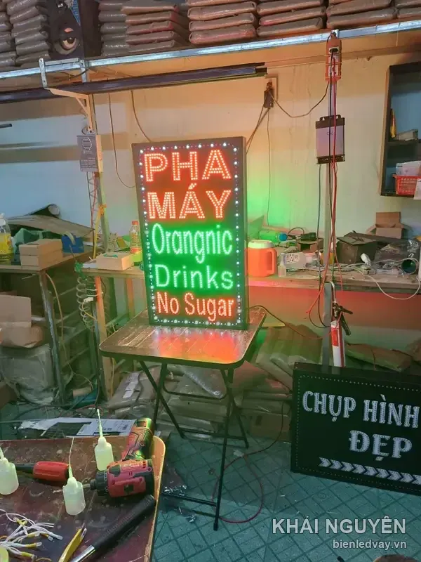Biển vẫy dọc viền LED bóng tròn thử sáng tại xưởng, có chữ đỏ PHA MÁY và chữ xanh lá Orangnic Drinks No Sugar.