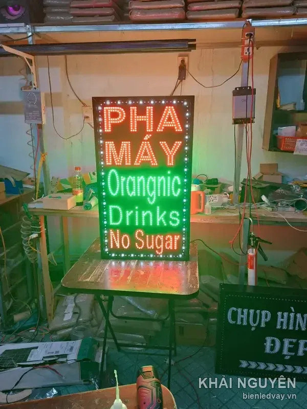 Ảnh cận biển vẫy PHA MÁY Orangnic Drinks No Sugar nền đen, viền bóng LED trắng, phía sau là bàn nguội của xưởng