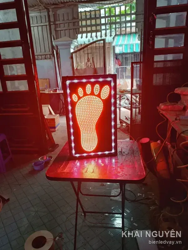 Biển vẫy hình bàn chân bóng LED vàng trên nền đỏ, viền bóng tròn trắng, đặt trên bàn xếp mặt đỏ giữa xưởng