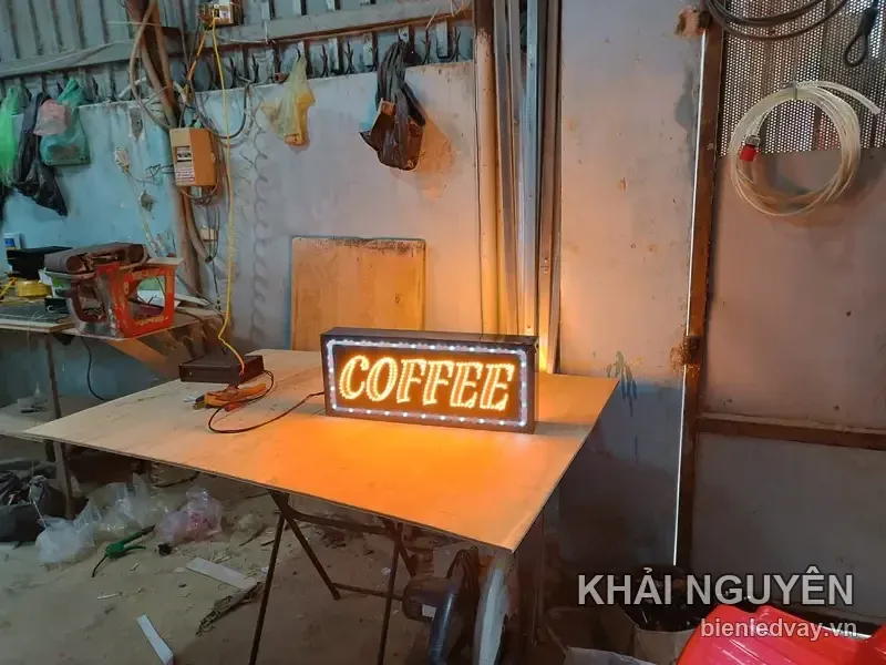 Biển vẫy COFFEE bóng LED vàng cam đang được đấu nguồn thử sáng tại xưởng, hắt vệt sáng ấm xuống mặt bàn gỗ.