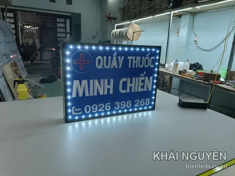 Biển vẫy Quầy Thuốc Minh Chiến nền xanh dương, viền bóng LED trắng nhấp nháy, đặt trên bàn xưởng