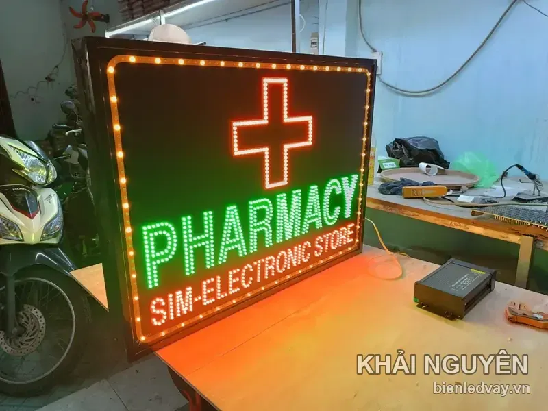 Biển vẫy PHARMACY SIM-ELECTRONIC STORE chụp góc nghiêng, chữ thập LED đỏ, viền bóng vàng, cạnh bộ nguồn LED