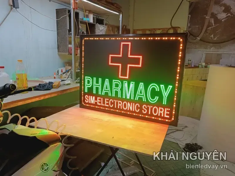 Biển PHARMACY SIM-ELECTRONIC STORE chữ xanh lá trên nền đen, chữ thập LED đỏ, viền bóng vàng, khung gỗ sẫm