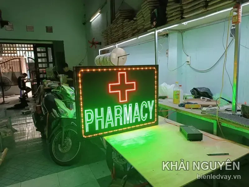 Biển vẫy nhà thuốc chữ PHARMACY xanh lá và dấu thập đỏ trên nền đen, viền LED bóng tròn vàng, thử sáng tại xưởng