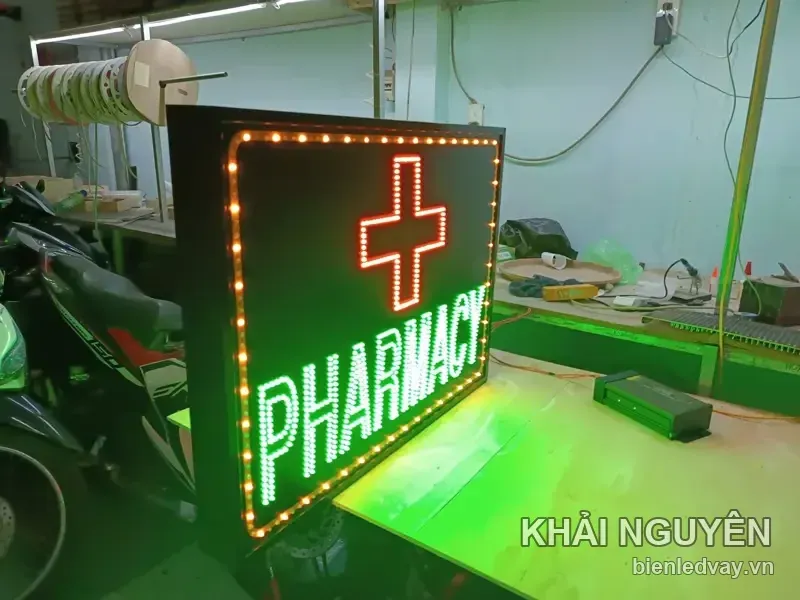 Ảnh nghiêng biển vẫy PHARMACY cho thấy thùng đen dày, dấu thập đỏ và hàng LED bóng tròn vàng chạy quanh viền