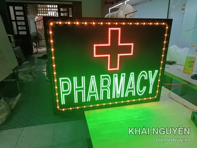 Cận cảnh biển vẫy PHARMACY chính diện, dấu thập đỏ nổi trên nền đen, ánh xanh lá hắt xuống mặt bàn gỗ