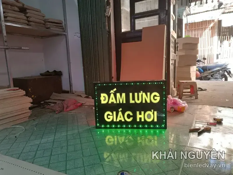 Biển vẫy chữ LED vàng ĐẤM LƯNG – GIÁC HƠI trên nền đen, viền bóng tròn xanh lá, đặt dưới nền gạch trước cửa xưởng