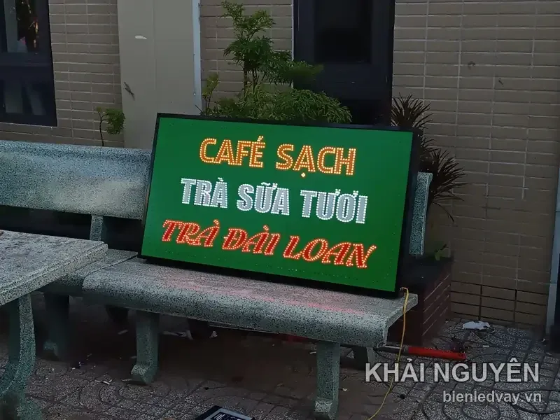 Biển vẫy CAFÉ SẠCH - TRÀ SỮA TƯƠI - TRÀ ĐÀI LOAN nền xanh lá, góc chụp nghiêng lúc chạng vạng, dây nguồn đang cắm thử.