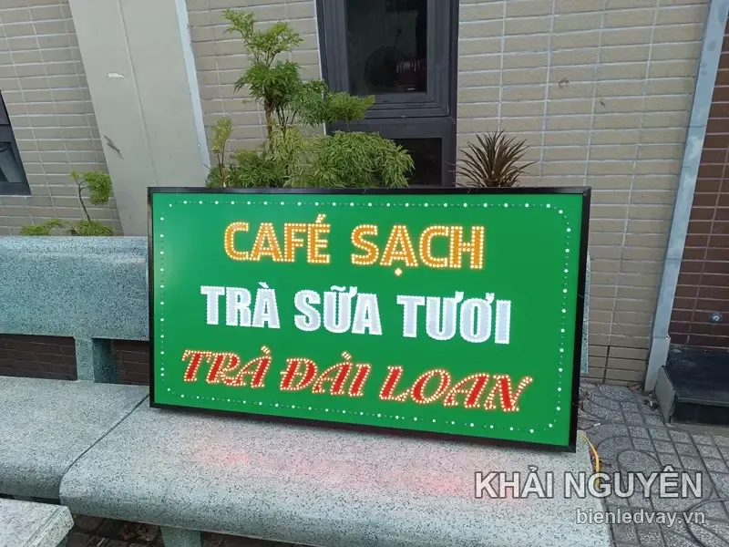 Biển vẫy nền xanh lá viền bóng LED trắng, chữ CAFÉ SẠCH màu vàng nổi bật, đặt tựa ghế đá trước bức tường gạch.