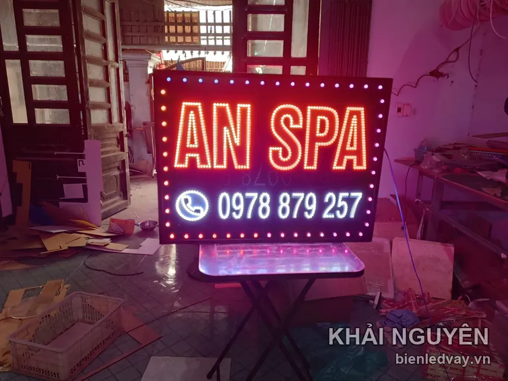 Biển vẫy An Spa cỡ lớn, chữ đỏ viền cam rực, số liên hệ trên biển trắng, viền bóng LED nhiều màu, đặt trên bàn xếp