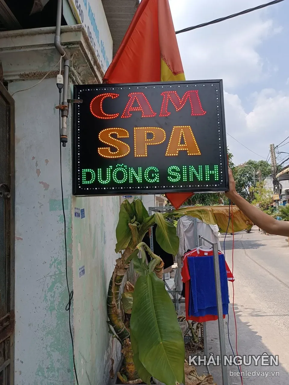 Biển vẫy LED Cam Spa dưỡng sinh vừa lắp lên tường vôi cũ ngoài phố, thợ đưa tay đỡ, chữ đỏ vàng xanh trên nền đen