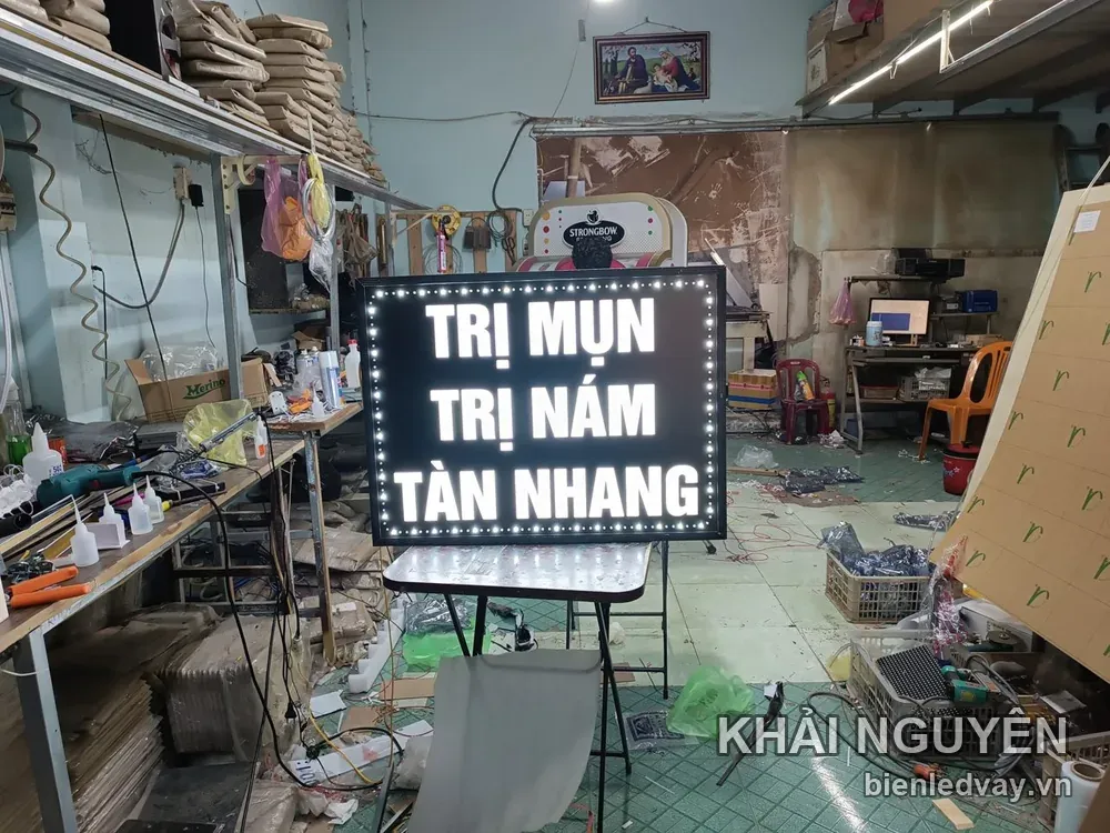 Biển vẫy nền đen chữ trắng TRỊ MỤN – TRỊ NÁM – TÀN NHANG, viền LED bóng tròn trắng, dựng trên giá tại xưởng