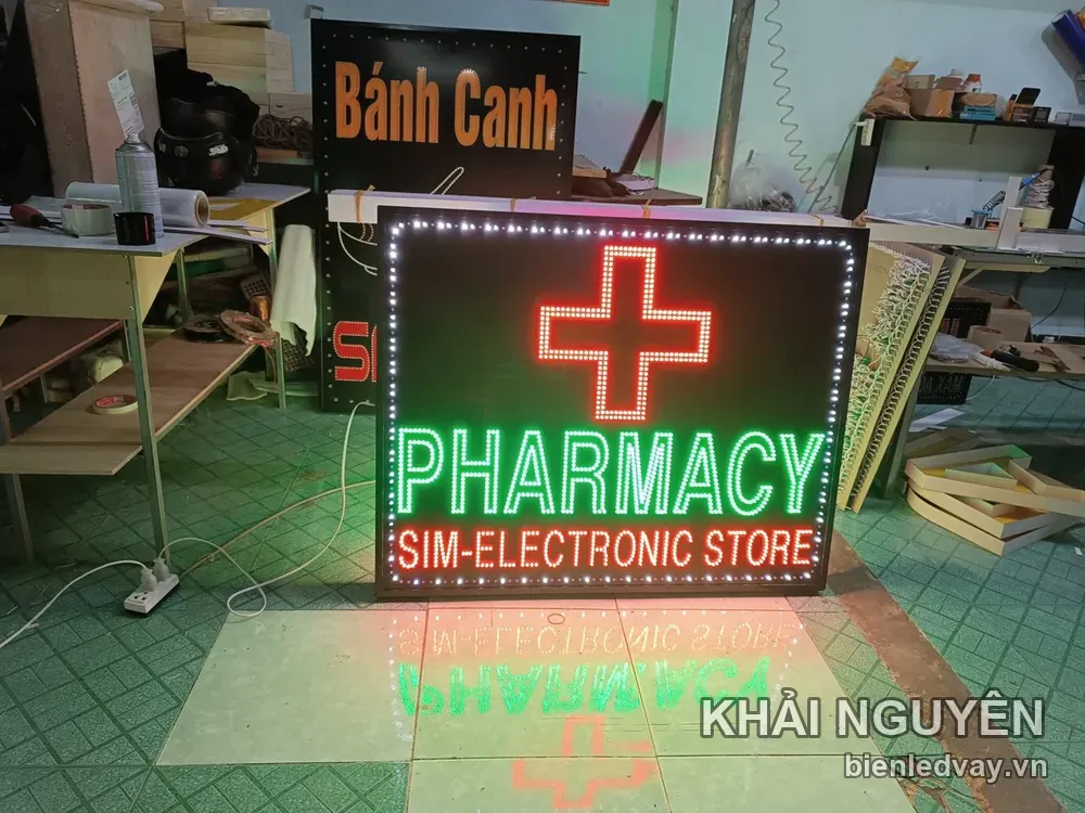 Biển vẫy PHARMACY có nền đen, chữ xanh lá, dòng SIM-ELECTRONIC STORE, chữ thập đỏ và viền bóng trắng.