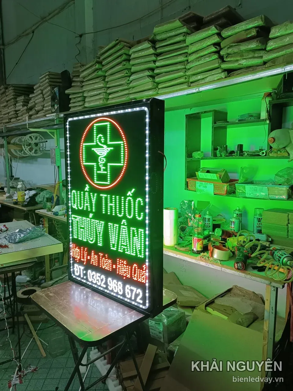 Biển vẫy Quầy Thuốc Thúy Vân được dựng nghiêng trên bàn xếp giữa xưởng, với chữ LED xanh lá và chữ thập đỏ viền tròn.