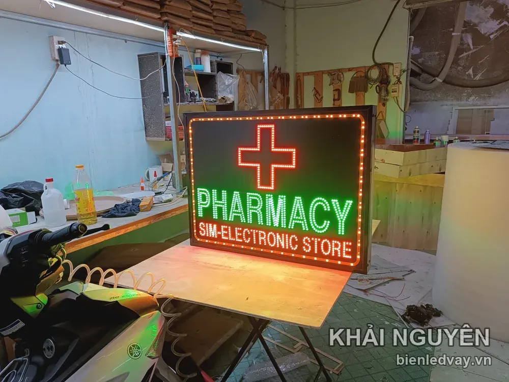 Biển vẫy PHARMACY có chữ xanh lá, dòng SIM-ELECTRONIC STORE, chữ thập LED đỏ, viền bóng tròn cam và khung đen.