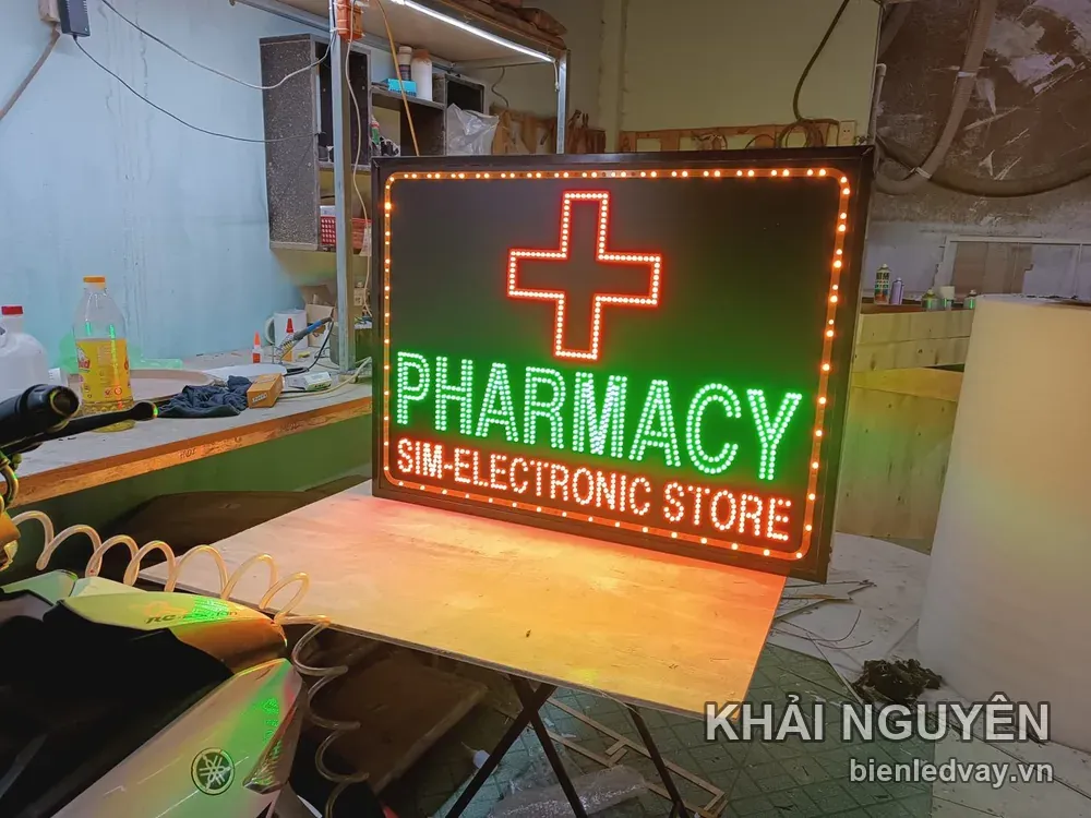 Biển vẫy PHARMACY - SIM-ELECTRONIC STORE có viền bóng LED vàng cam, chữ thập đỏ và đặt trên bàn gỗ ở xưởng