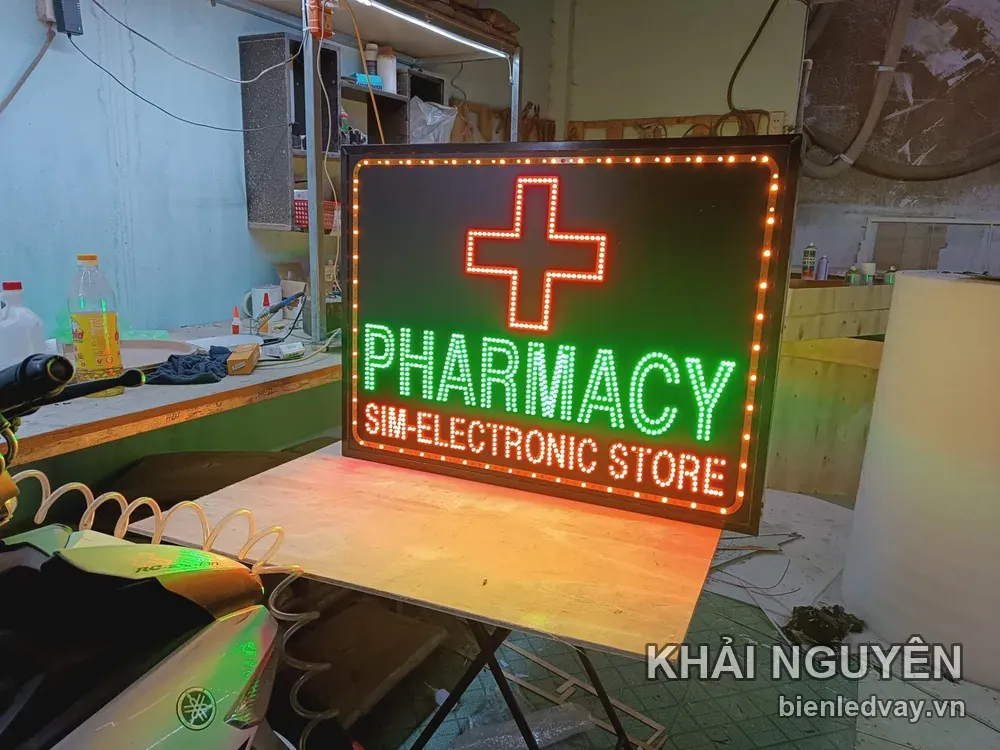 Biển vẫy PHARMACY - SIM ELECTRONIC STORE đặt trên bàn gỗ, có nền đen, chữ xanh lá, chữ thập và viền LED màu cam.