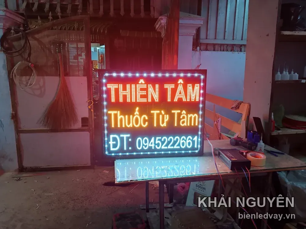 Ảnh cận biển THIÊN TÂM Thuốc Từ Tâm trên bàn gỗ xưởng, hai hàng chữ cam, viền LED trắng, bóng hắt xuống mặt bàn