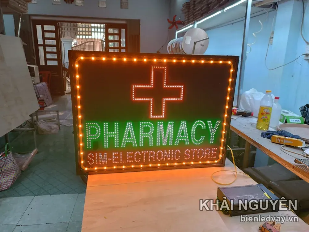 Biển vẫy nền đen có chữ PHARMACY xanh lá, dòng SIM-ELECTRONIC STORE, chữ thập LED đỏ trắng và viền LED vàng.