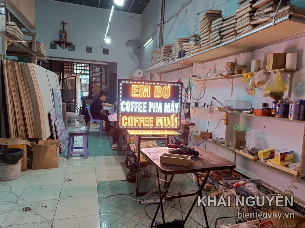 Biển nền đen viền bóng LED trắng đặt trong xưởng, gồm chữ LED cam EM BƠ và hai dòng COFFEE PHA MÁY, COFFEE MUỐI.