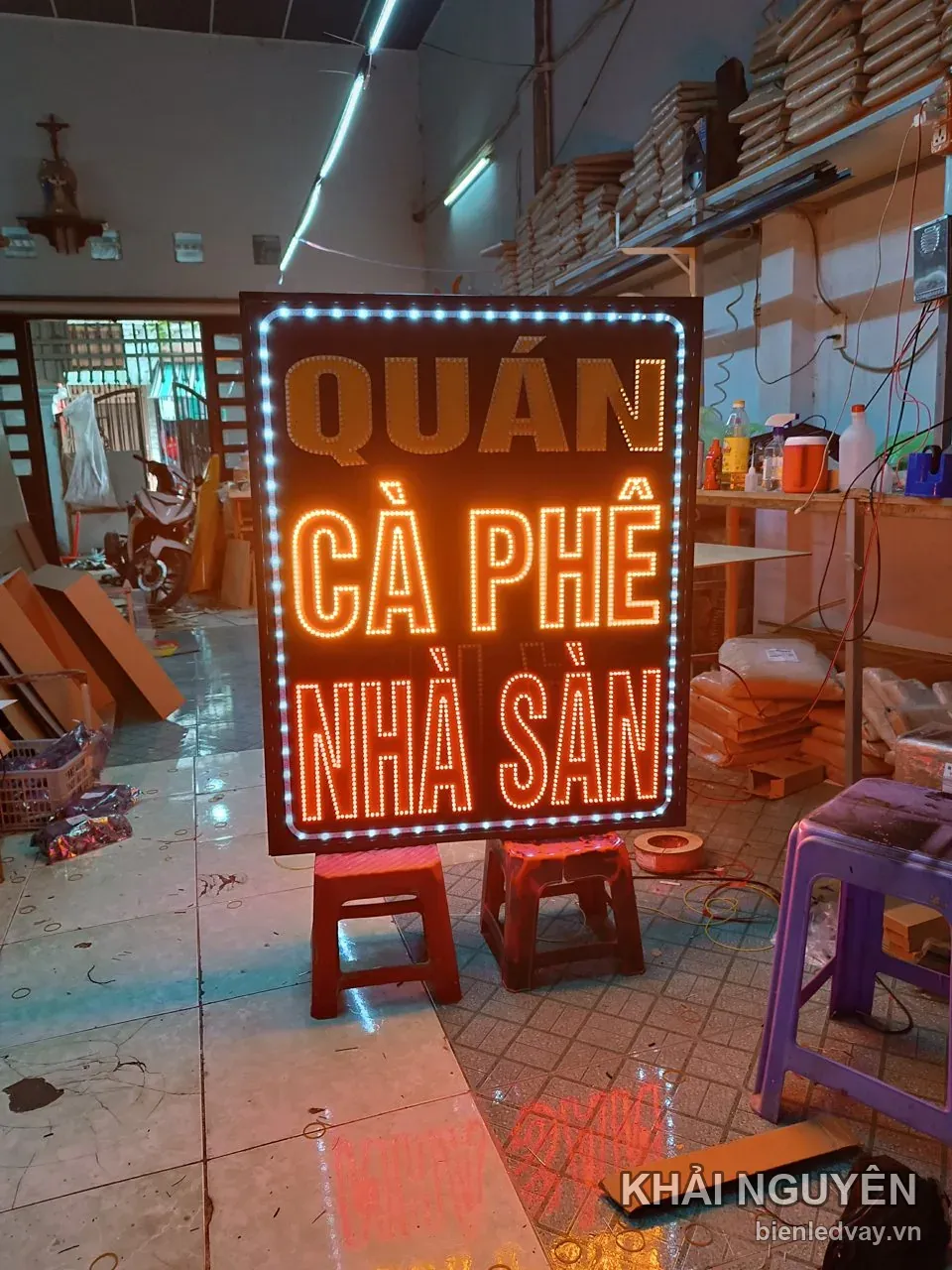 Biển vẫy LED Quán Cà Phê Nhà Sàn thiết kế khổ đứng, chữ vàng cam nền đen, viền LED bóng tròn trắng, được kê trên ghế nhựa đỏ