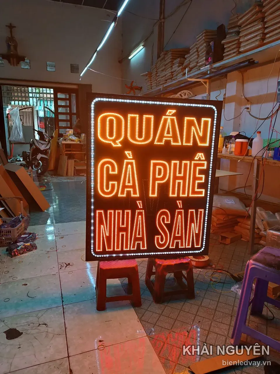Biển QUÁN CÀ PHÊ NHÀ SÀN có ba dòng chữ LED cam trên nền nâu sẫm, viền bóng LED trắng, đang kê trên ghế nhựa ở xưởng.