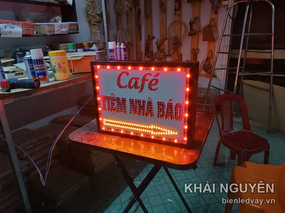 Biển Café Tiệm Nhà Báo với chữ đỏ trên nền trắng, viền bóng đèn tròn đỏ và dải LED vàng chạy dưới chân biển.
